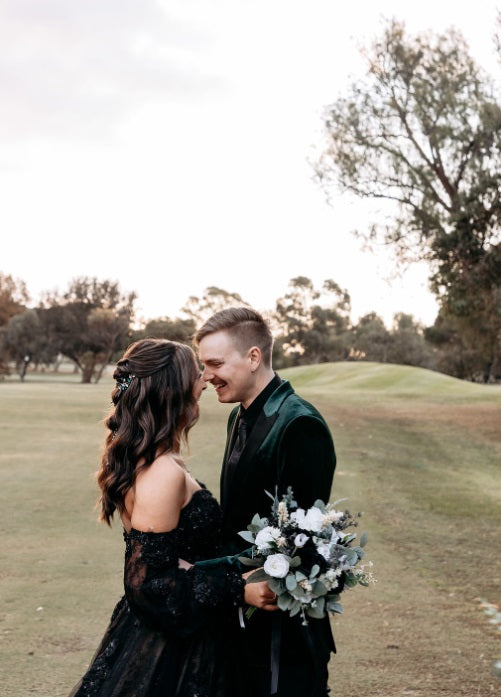 The bride wears a black off-the-shoulder lace dress and holds a bouquet of faux white and green flowers. Flowers by Flowers For Ever After