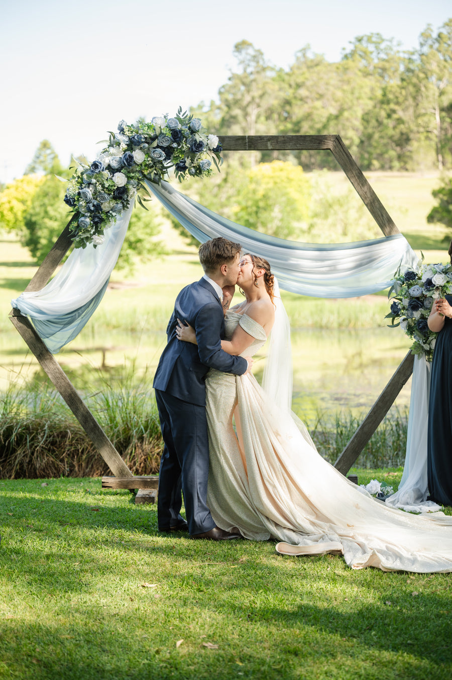 A bride in a cream off-the-shoulder wedding gown and a groom in a navy suit embrace and kiss under a rustic hexagonal wooden arch decorated with artificial blue and white flowers.Flowers by Flowers For Ever After
