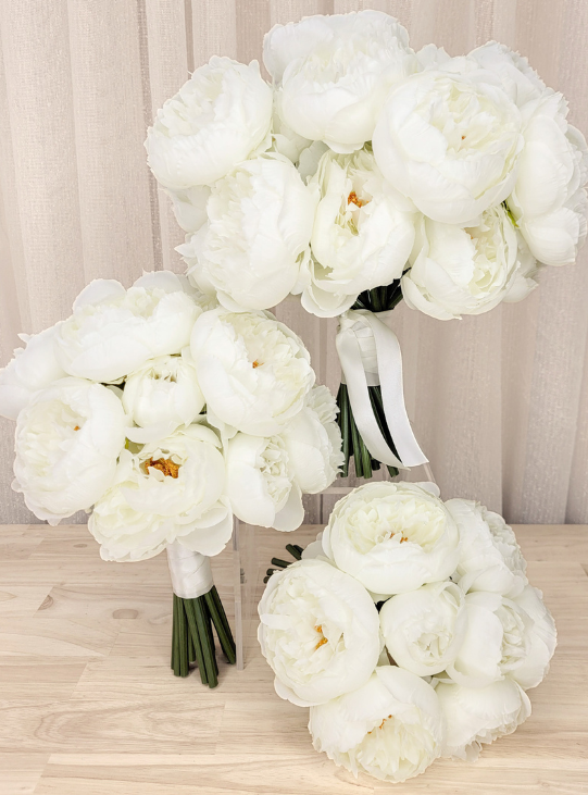Faux white peony bouquets with green stems and white ribbon wraps, arranged on a light wood surface against a beige curtain backdrop. Made in Melbourne by Flowers For Ever After, wedding flower packages shipping worldwide, custom orders available