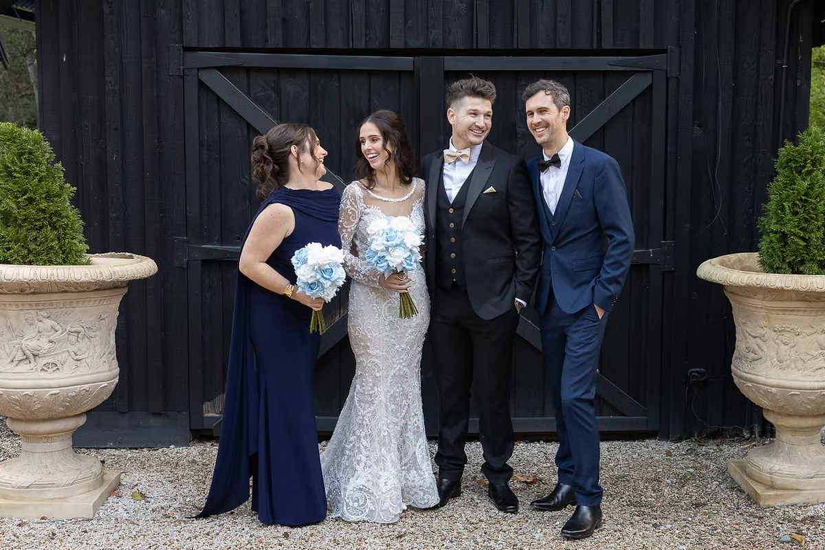 The central couple wears wedding attire, with the bride in a white lace gown holding a light blue and white bouquet and the groom in a black suit with gold accents. Flowers by Flowers For Ever After