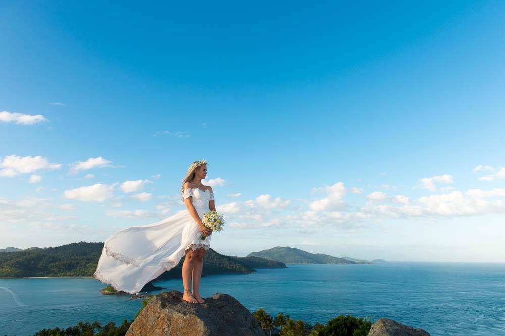 A bride in a flowing white wedding dress stands on a rocky cliff overlooking a blue ocean and distant green islands under a bright sky. Holding a bouquet of faux daisies. Flowers by Flowers For Ever After