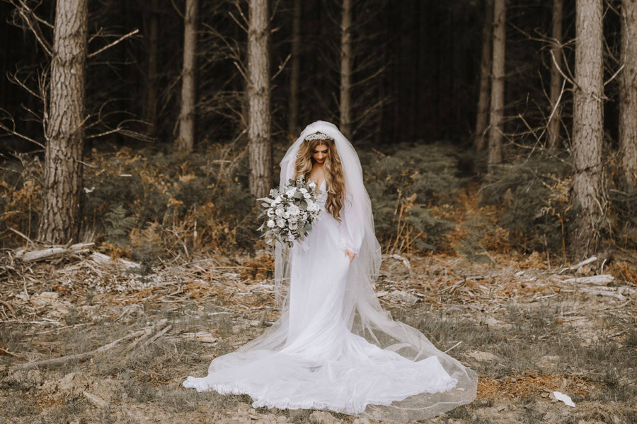 A bride in a long white wedding gown and sheer veil stands in a forest clearing holding a bouquet of white faux flowers. Flowers by Flowers For Ever After