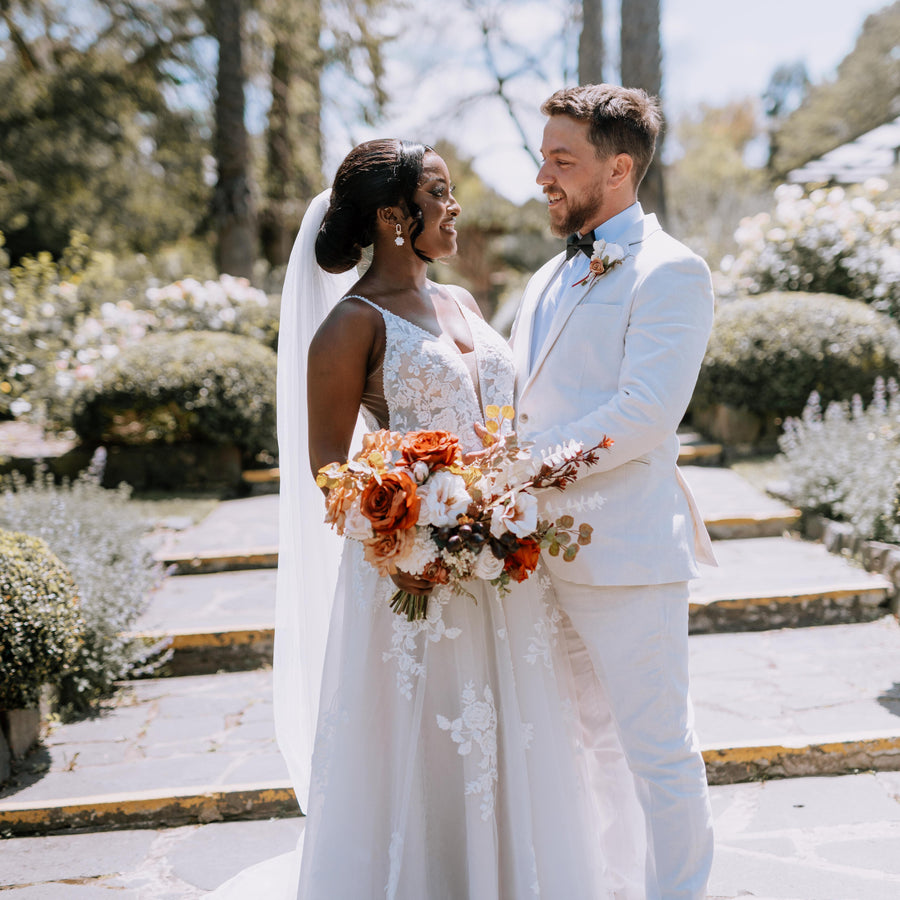 A bride in a white lace wedding gown with a long veil and a groom in a white suit holding a large orange and white artificial floral bouquet, standing on stone steps in a garden setting. Flowers by Flowers For Ever After