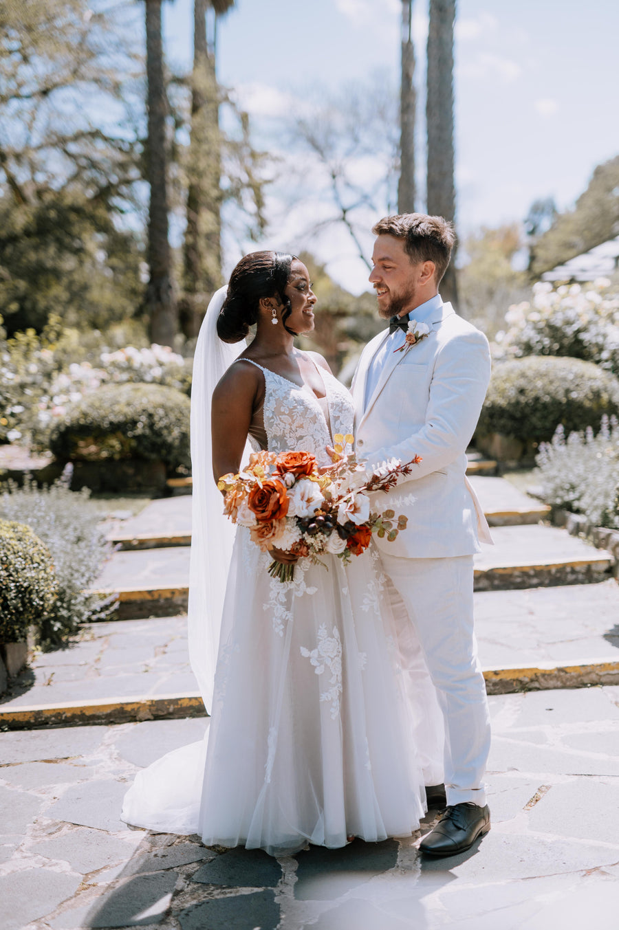 A bride in a white lace wedding gown with a long veil and a groom in a white suit holding a large orange and white artificial floral bouquet, standing on stone steps in a garden setting. Flowers by Flowers For Ever After