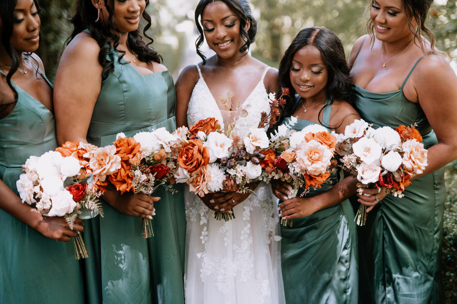 A bride in a white lace wedding dress stands with four bridesmaids wearing matching sage green satin dresses, each holding a large bouquet of white, orange, and burgundy flowers, photographed outdoors in soft natural light. Flowers by Flowers For Ever After