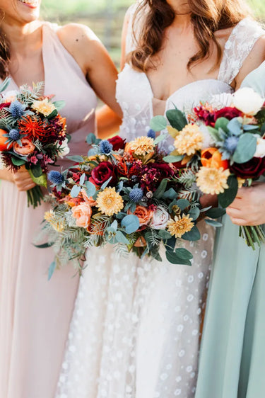 Three bridesmaids holding large, colorful faux wildflower bouquets. The bride in the center wears a white lace dress, flanked by bridesmaids in blush pink and sage green dresses, with a softly blurred outdoor background. Flowers by Flowers For Ever After