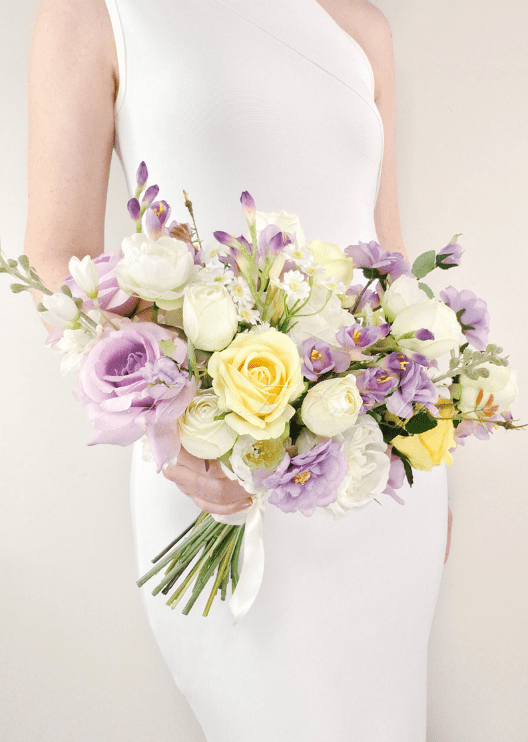 Artificial wedding bridal bouquet held by a bride, with lilac faux roses, yellow roses, white peonies and purple flower spikes. Made in Melbourne by Flowers For Ever After, shipping Australia and Worldwide.