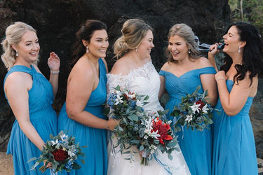 Five women posing outdoors in front of a dark rock face, featuring four bridesmaids in matching bright blue off-the-shoulder dresses and a bride in a white lace wedding gown, each holding a floral bouquet. Flowers by Flowers For Ever After