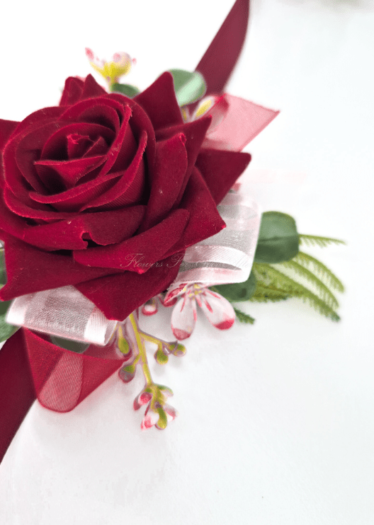 Artificial wedding corsage close-up showing burgundy faux rose petals, tiny pink blossoms, green foliage and white and burgundy ribbon loops. For formals, proms and ball occasions. Made in Melbourne by Flowers For Ever After, shipping Australia and Worldwide.