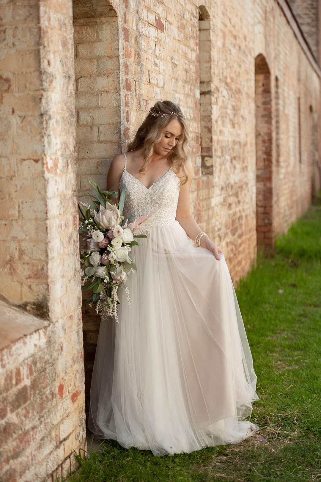 A bride in a white tulle wedding dress with a beaded bodice leans against a rustic brick wall, holding a large faux floral bridal bouquet. Flowers by Flowers For Ever After