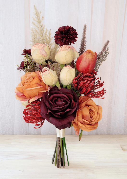 Artificial wedding bouquet with orange and burgundy faux roses, cream-pink rosebuds, red spiky flowers, burgundy chrysanthemum and pale plumes. Made in Melbourne by Flowers For Ever After, shipping Australia and Worldwide.