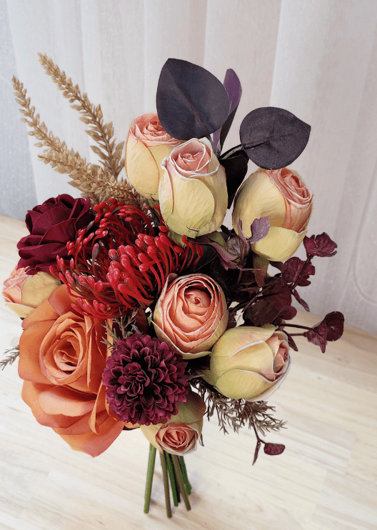 Artificial wedding small bouquet, overhead view of orange and burgundy faux roses, cream-pink rosebuds, red spiky bloom and dark leaves. Made in Melbourne by Flowers For Ever After, shipping Australia and Worldwide.