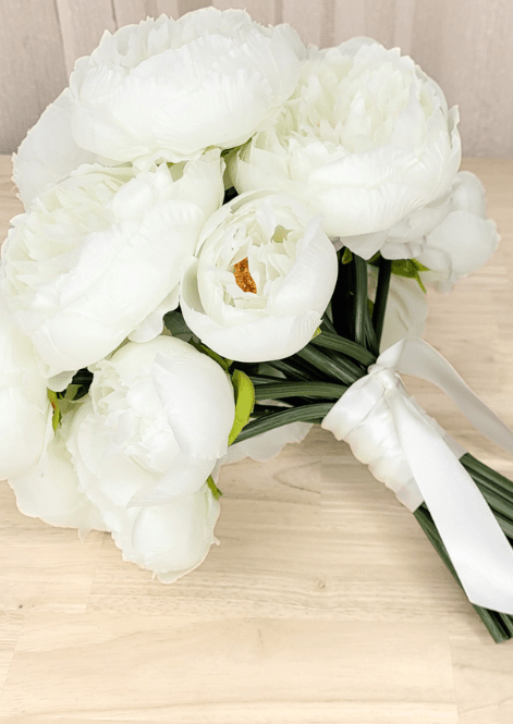 Artificial wedding bouquet laid sideways, showing white faux peonies, golden flower centres, green stems and white ribbon tails. Made in Melbourne by Flowers For Ever After, shipping Australia and Worldwide.