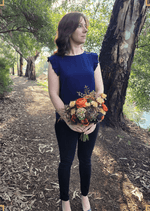 Artificial wedding bridal bouquet held in both hands, featuring orange, yellow and burgundy faux blooms with green foliage. Made in Melbourne by Flowers For Ever After, shipping Australia and Worldwide.