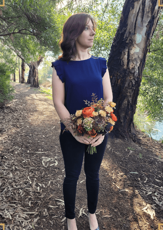 Artificial wedding bridal bouquet held in both hands, featuring orange, yellow and burgundy faux blooms with green foliage. Made in Melbourne by Flowers For Ever After, shipping Australia and Worldwide.