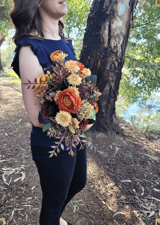 Artificial wedding bridal bouquet held by a bride, with orange and yellow faux flowers, burgundy accents and trailing ribbons. Made in Melbourne by Flowers For Ever After, shipping Australia and Worldwide.