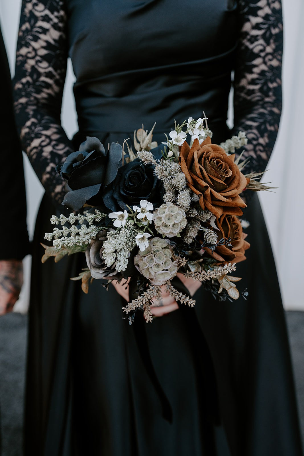 A close-up of a person in a black satin dress holding a gothic-style faux flower bouquet with black and rust-brown roses, white filler flowers, and muted greenery. Flowers by Flowers For Ever After