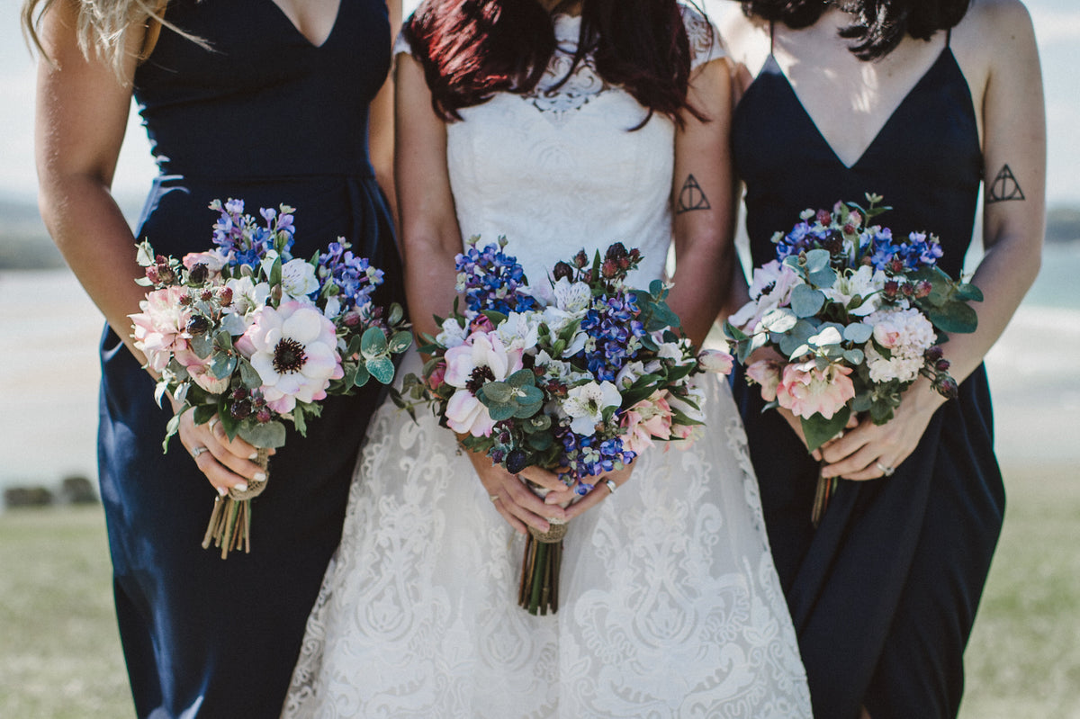Bride and two bridesmaids standing outdoors, each holding a bouquet of pink, blue, and white flowers. The bridesmaids wear dark navy sleeveless dresses, and the bride wears a white lace dress. Flowers by Flowers For Ever After