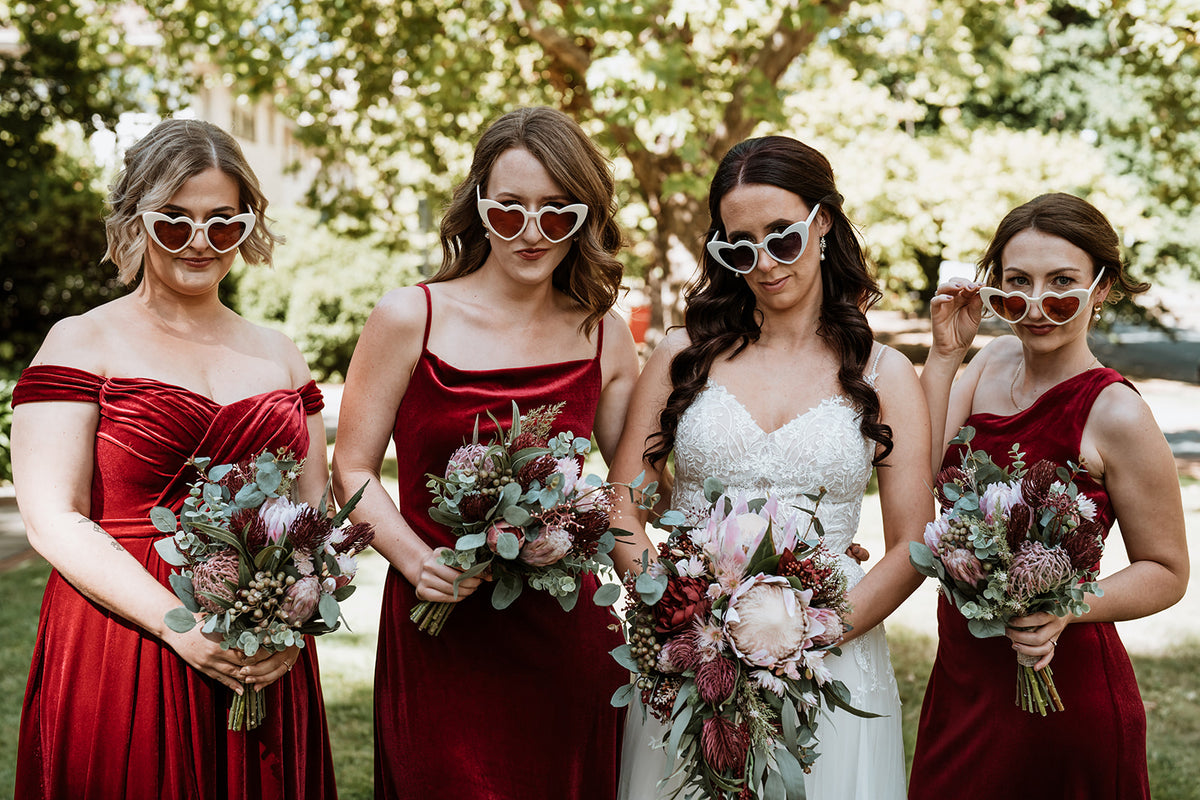 Four women posing outdoors in wedding attire, with three bridesmaids in deep red dresses and the bride in a white gown, each wearing heart-shaped sunglasses and holding  red and pink faux bouquets. Flowers by Flowers For Ever After