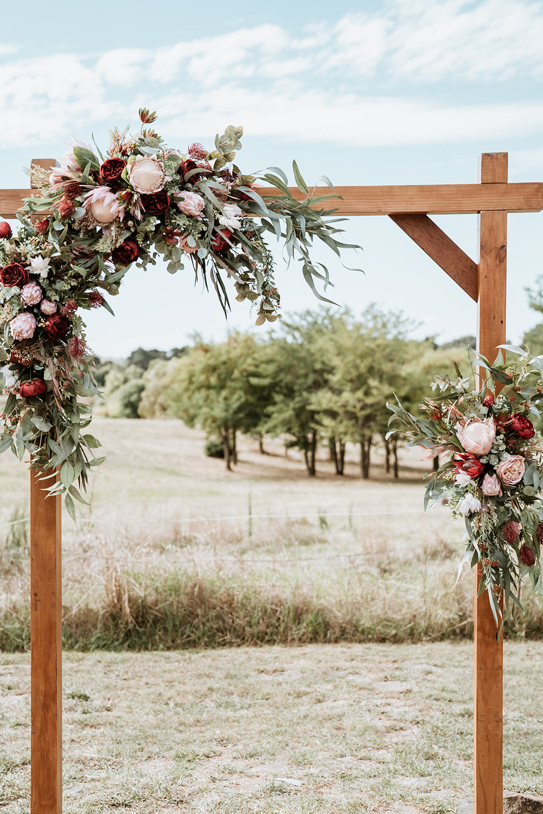 A rustic wooden archway decorated with lush faux floral arrangements of pink peonies, burgundy roses, and green eucalyptus, set against an outdoor grassy field with trees under a cloudy sky.Flowers by Flowers For Ever After