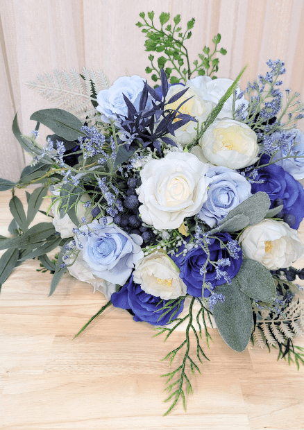 Artificial wedding bouquet laid sideways, showing navy and pale blue faux roses, white ranunculus, blue berries and eucalyptus. Made in Melbourne by Flowers For Ever After, shipping Australia and Worldwide.