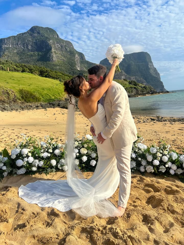 A bride in a white wedding gown with a long train and veil, and a groom in a light beige suit, kiss on a sandy beach with mountains and ocean in the background. Lord Howe Island.  Flowers by Flowers For Ever After