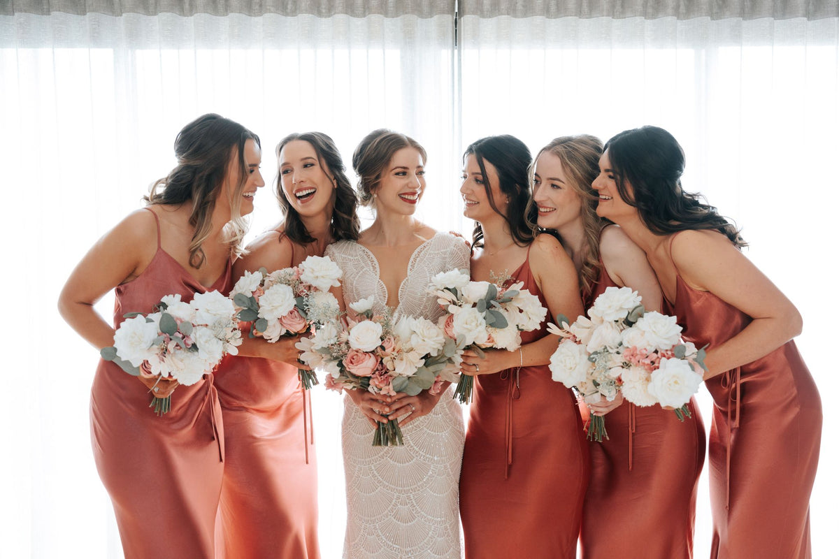 A bride in a white lace wedding gown stands with five bridesmaids wearing matching dusty rose satin dresses, holding white and blush artificial bouquets in front of a bright window with sheer curtains.