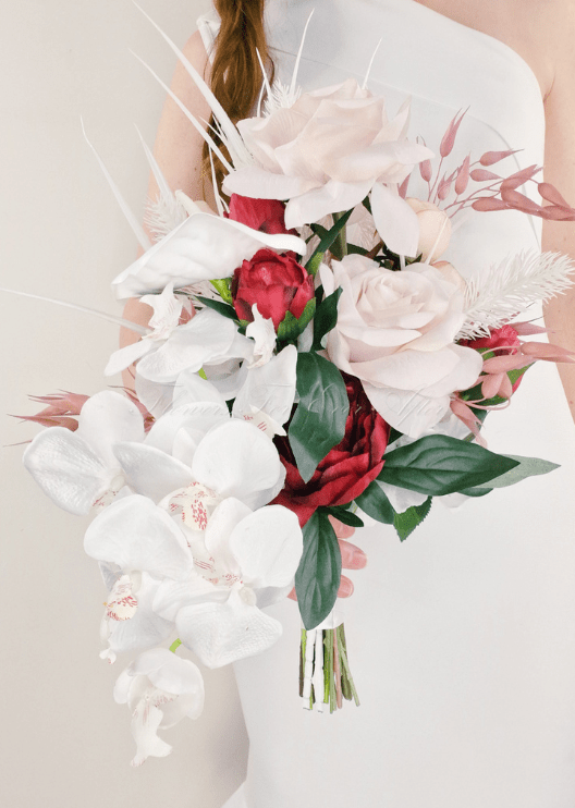 Artificial wedding cascading bouquet held by a bride, with blush faux roses, red blooms, white orchids, pink foliage and white feathery sprigs. Made in Melbourne by Flowers For Ever After, shipping Australia and Worldwide.