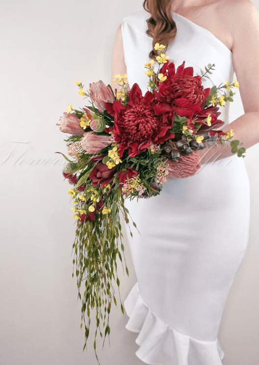Artificial wedding cascading bridal bouquet carried by a bride, with red faux waratah, pink protea, yellow blooms and eucalyptus. Made in Melbourne by Flowers For Ever After, shipping Australia and Worldwide.
