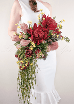 Artificial wedding cascading bridal bouquet held by a bride, with red faux waratah, pink protea, yellow blooms and long trailing foliage. Made in Melbourne by Flowers For Ever After, shipping Australia and Worldwide.
