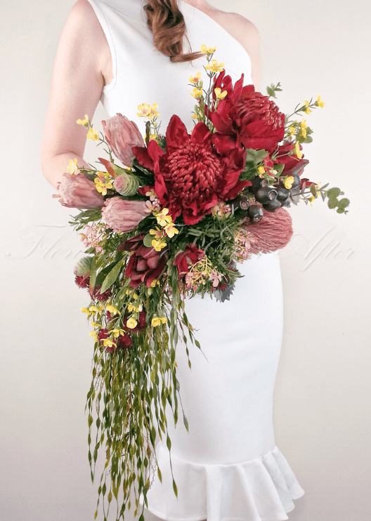 Artificial wedding cascading bridal bouquet held by a bride, with red faux waratah, pink protea, yellow blooms and long trailing foliage. Made in Melbourne by Flowers For Ever After, shipping Australia and Worldwide.