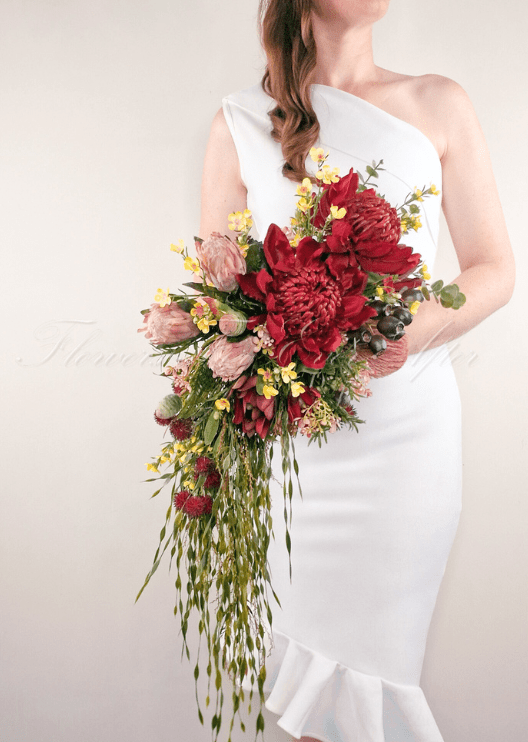 Artificial wedding cascading bridal bouquet held by a bride, with red faux waratah, pink protea, yellow flowers and long trailing foliage. Made in Melbourne by Flowers For Ever After, shipping Australia and Worldwide.