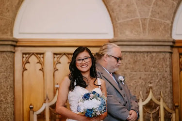 A smiling woman in a white wedding dress holding a blue and white bouquet stands next to a man in a gray suit in front of a stone archway and wooden altar.