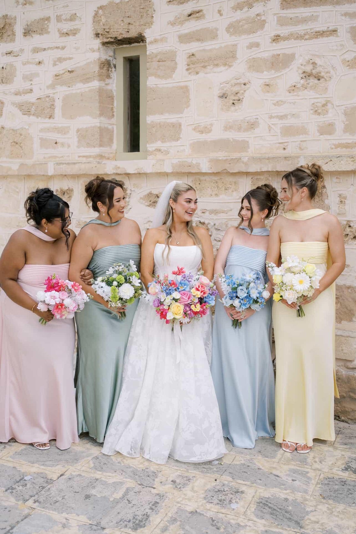A bride in a white lace wedding gown stands with five bridesmaids in pastel pink, sage green, light blue, and pale yellow dresses, holding colorful flower bouquets against a rustic stone wall. Flowers by Flowers For Ever After
