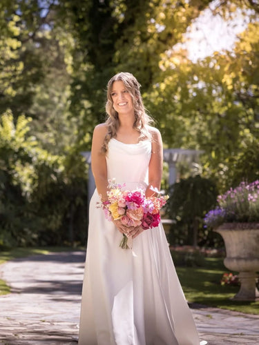 A woman wearing a sleeveless white wedding gown stands outdoors on a stone path holding a colorful bouquet of pink, red, and yellow flowers. Flowers by Flowers For Ever After