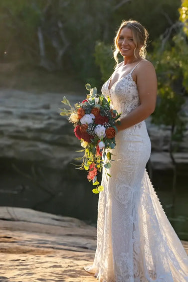 A woman wearing a sleeveless lace wedding gown stands outdoors by a rocky body of water, holding a large floral bouquet with red, white, and green flowers.Flowers by Flowers For Ever After