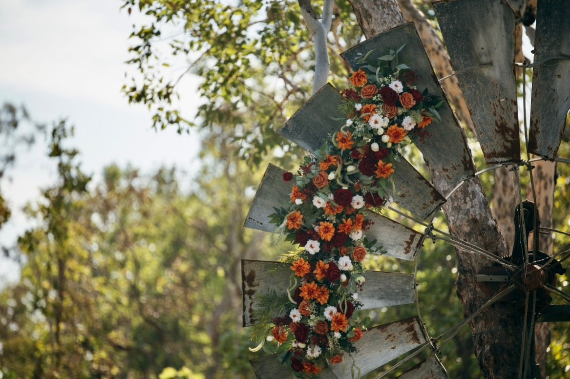 A rustic floral arrangement in orange, burgundy, and white flowers with green foliage, displayed on weathered metal windmill blades outdoors against a blurred forest background. Flowers by Flowers For Ever After