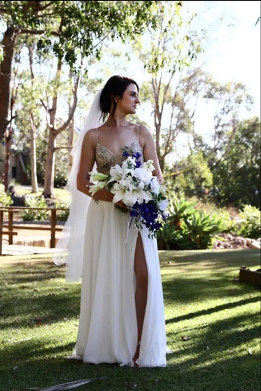 A woman wearing a sleeveless white wedding dress with a high slit and holding a bouquet of white and purple flowers, standing outdoors on a grassy lawn with trees in the background. Flowers by Flowers For Ever After