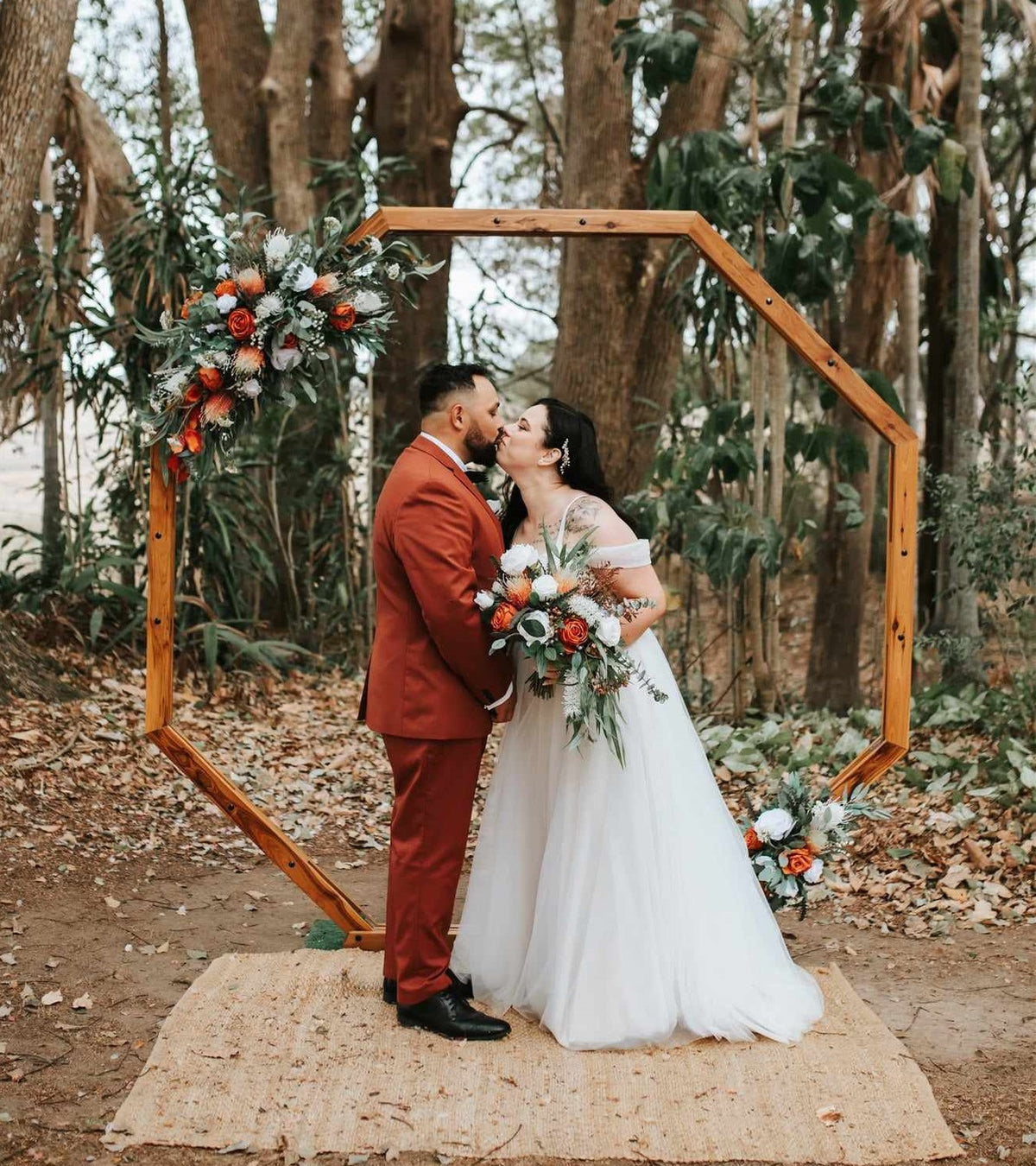 A bride in a white tulle wedding gown and a groom in a rust-brown suit kiss under a wooden hexagonal arch decorated with artificial orange and white flowers, set in a wooded outdoor venue. Flowers by Flowers For Ever After