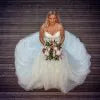A woman wearing a voluminous white wedding dress with a sheer tulle overlay and holding a bouquet, standing against a dark wood plank background.