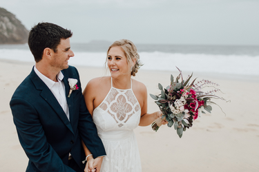 A bride in a white lace halter wedding dress holding a burgundy and green faux floral bouquet, standing on a sandy beach with the ocean and overcast sky in the background, next to a groom in a navy suit. Flowers by Flowers For Ever After