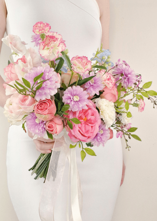 Artificial wedding bridal bouquet held by a bride, with pink faux roses, lilac daisies, white flowers and trailing ivory ribbons. Made in Melbourne by Flowers For Ever After, shipping Australia and Worldwide.