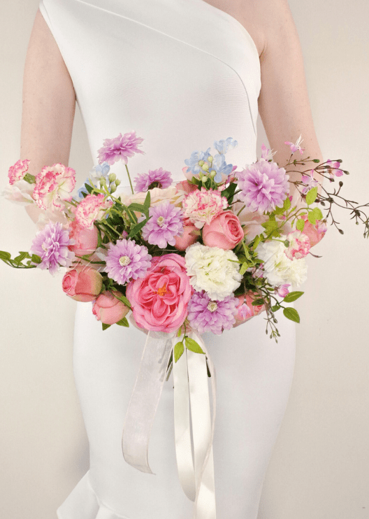 Artificial wedding bridal bouquet held by a bride, with pink faux roses, lilac daisies, white flowers, pale blue sprigs and ivory ribbons. Made in Melbourne by Flowers For Ever After, shipping Australia and Worldwide.