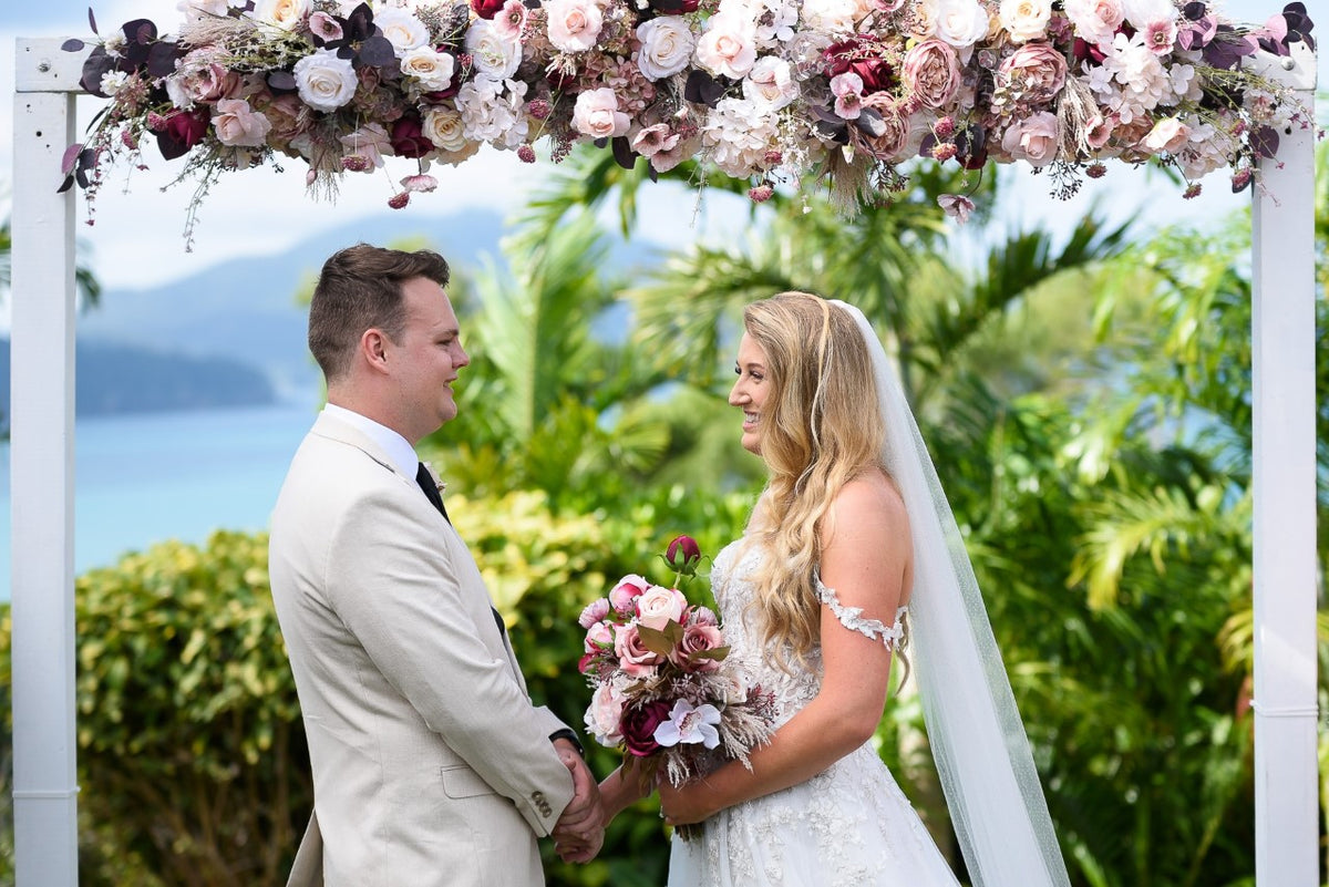 A bride in a white lace wedding dress and veil holding a artificial pink and white floral bouquet, standing under a white arch decorated with pink and white flowers, next to a groom in a light beige suit. Flowers by Flowers For Ever After