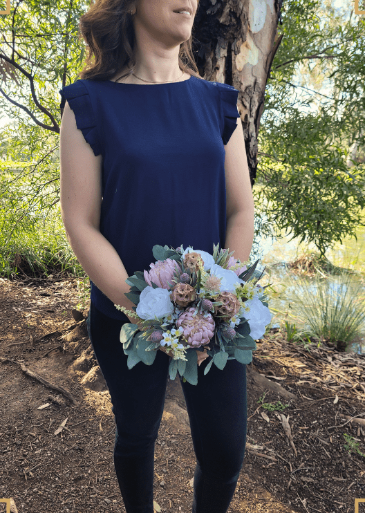 Artificial wedding bouquet held at waist height by a person, with ivory faux roses, blush pink proteas, tan lotus pods and eucalyptus. Made in Melbourne by Flowers For Ever After, shipping Australia and Worldwide.