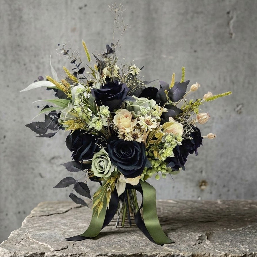 An artificial flower bouquet with green and black roses, muted green foliage, cream daisies, and olive green ribbons, displayed on a rough stone surface against a textured gray concrete wall. Made in Melbourne by  Flowers For Ever After, shipping Australia wide.