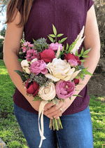 Artificial wedding bouquet held at waist height, with burgundy and ivory faux roses, mauve blooms, blue-grey berries and pale plumes. Made in Melbourne by Flowers For Ever After, shipping Australia and Worldwide.