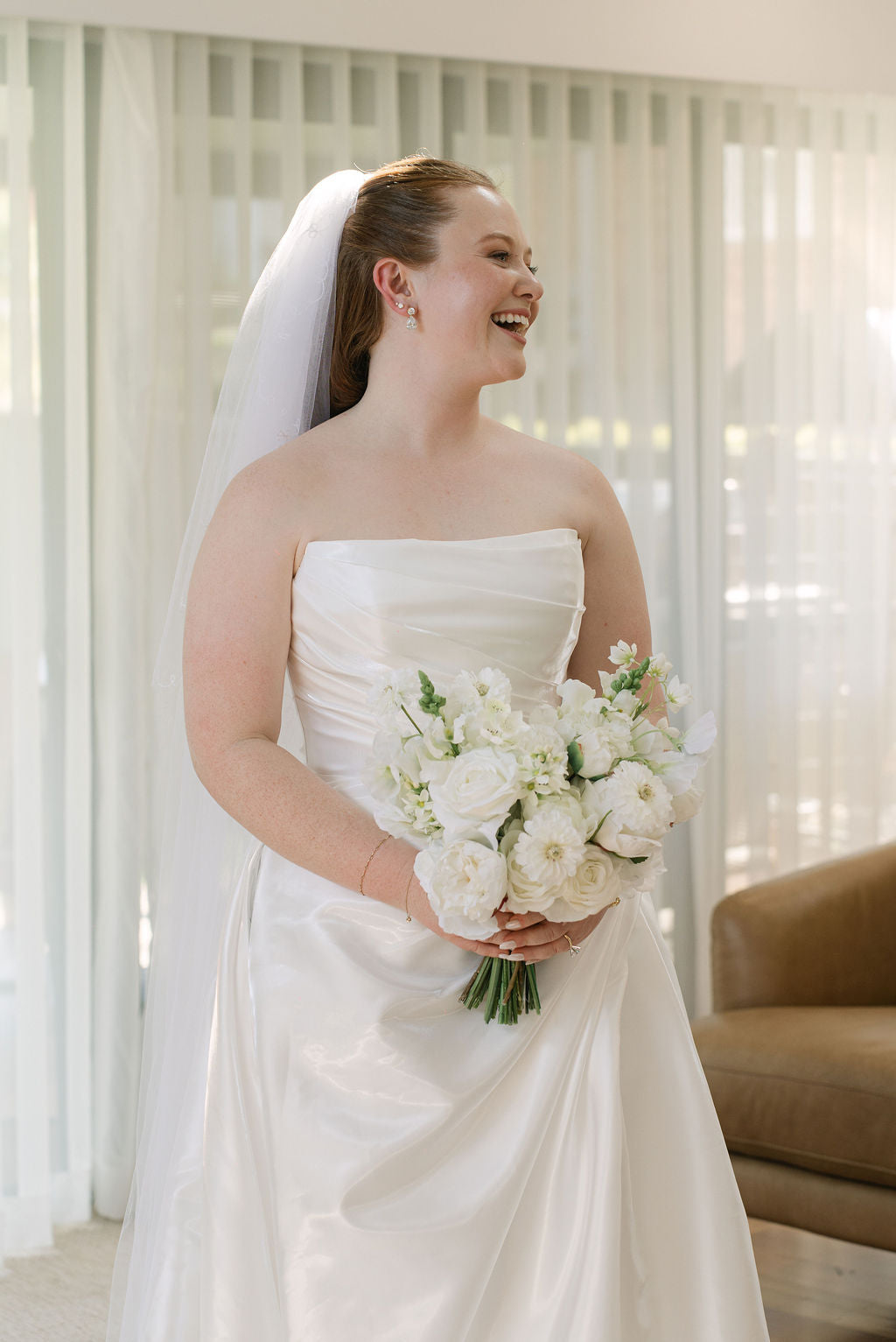 A woman wearing a strapless white satin wedding gown and long veil, holding a bouquet of white faux flowers. Flowers by Flowers For Ever After