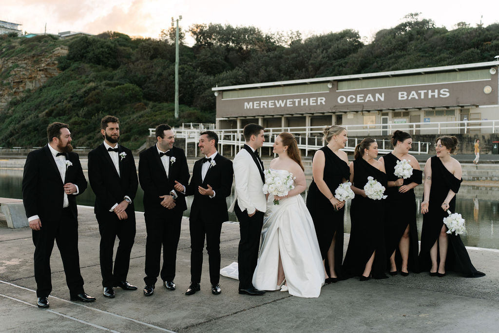 A wedding party stands outdoors in front of Merewether Ocean Baths. Bride and bridesmaids holding faux white wedding flower bouquets. Flowers by Flowers For Ever After