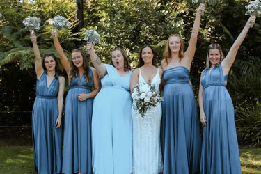 A bride in a white lace wedding gown holding a bouquet stands outdoors with five bridesmaids wearing matching dusty blue sleeveless maxi dresses, all raising their arms in celebration.